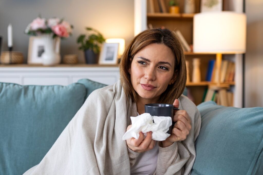Woman wrapped in a blanket on a sofa holding tissues and a mug